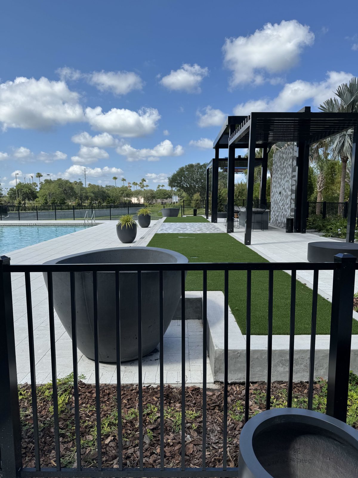Large charcoal concrete planters set on a pool deck beside a pergola and turf lawn at The Maxwell at Xentury City